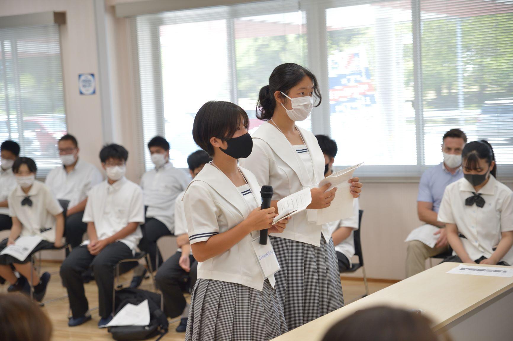オンライン交流会にて、カメラに向かって話をする県立由利高校の女子生徒2名の写真。1人は手持ちマイクを手にしており、もう1人は資料を持っている様子が確認できる
