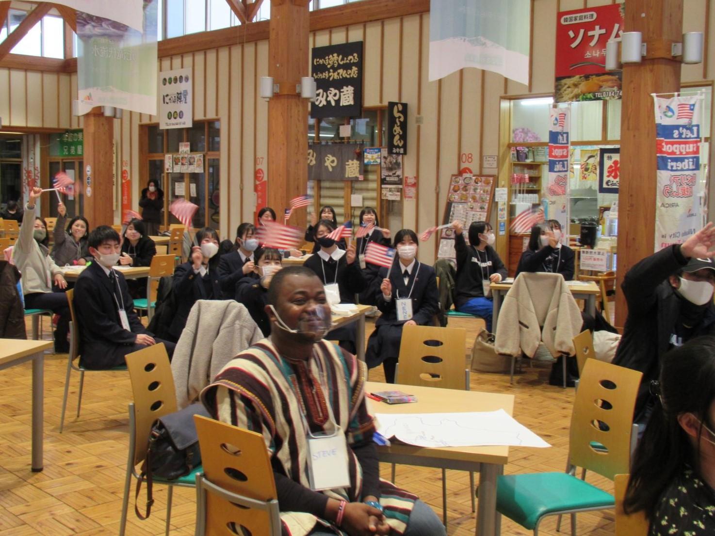 にかほ市観光拠点センター内のフードコートで、各々席に座ってミニサイズのリベリア国旗を振る参加者たちの写真。写真手前の席にはスティーブンス・モール氏が座っている様子も確認できる