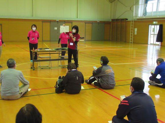 体育館の床に座ってインストラクターの説明を聞く参加者たちの写真。参加者たちの手元には各々運動量測定機器を持っている様子が確認できる