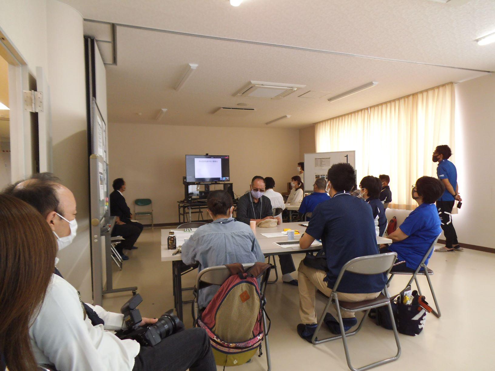 にかほ市スポーツ宿泊研修センターの会議室で、大型モニターを介して説明を受ける参加者たちの写真。モニターの画面には「みんなが生きやすい環境は」と書かれたテキストも確認できる