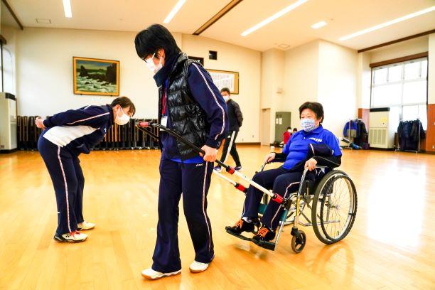 公民館館内で、取り付け式牽引補助具を付けた車イスを実際に引っ張てみる参加者の写真