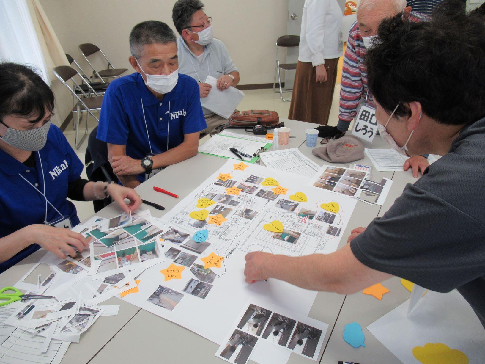 会議室における、象潟地区周辺のバリアフリーマップ作成風景の写真。大きな紙に印刷された地図に自分たちが撮影してきた施設内外各所の小さな写真を貼り、更にコメントが書かれた付箋を貼って行く様子が確認できる