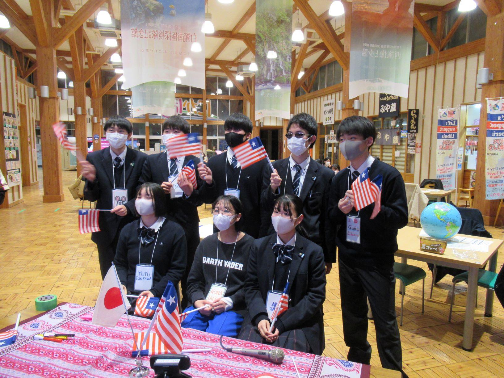 オンライン交流会にて、リベリアの応援スタイルを披露する仁賀保高校の生徒たち8人の写真。全員がミニサイズのリベリア国旗を持ち、サングラスをかけている生徒も確認できる
