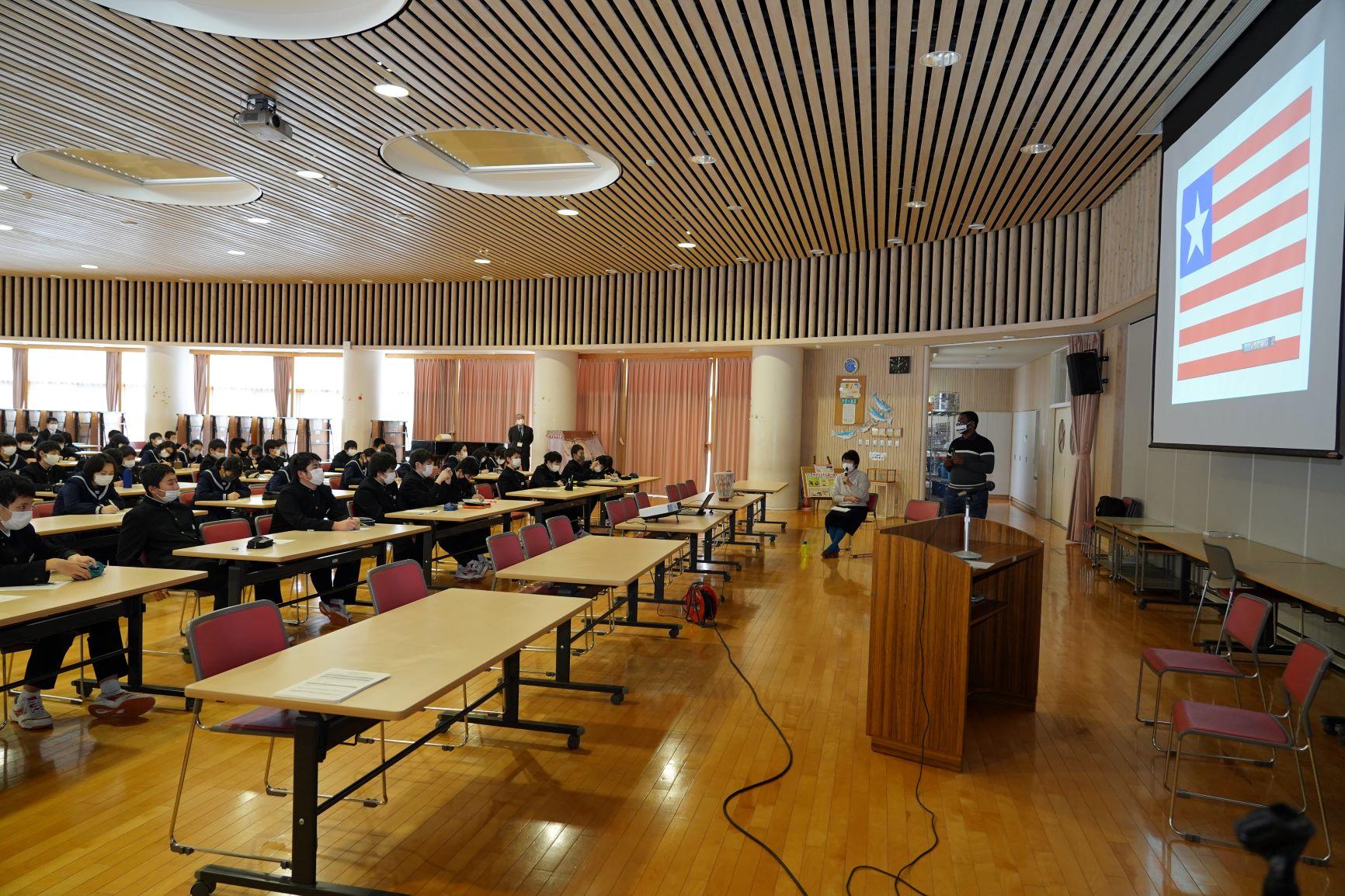演台の脇から撮影された、象潟中学校のランチルームにおけるリベリア講座の実施風景の写真。スクリーンにはリベリアの国旗が映し出されており、それに解説を行うスティーブンス・モール氏の姿が確認できる