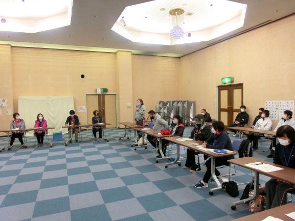 会議室にて、インストラクターを囲む様に並べられた長机に座って講義を受ける参加者たちの写真