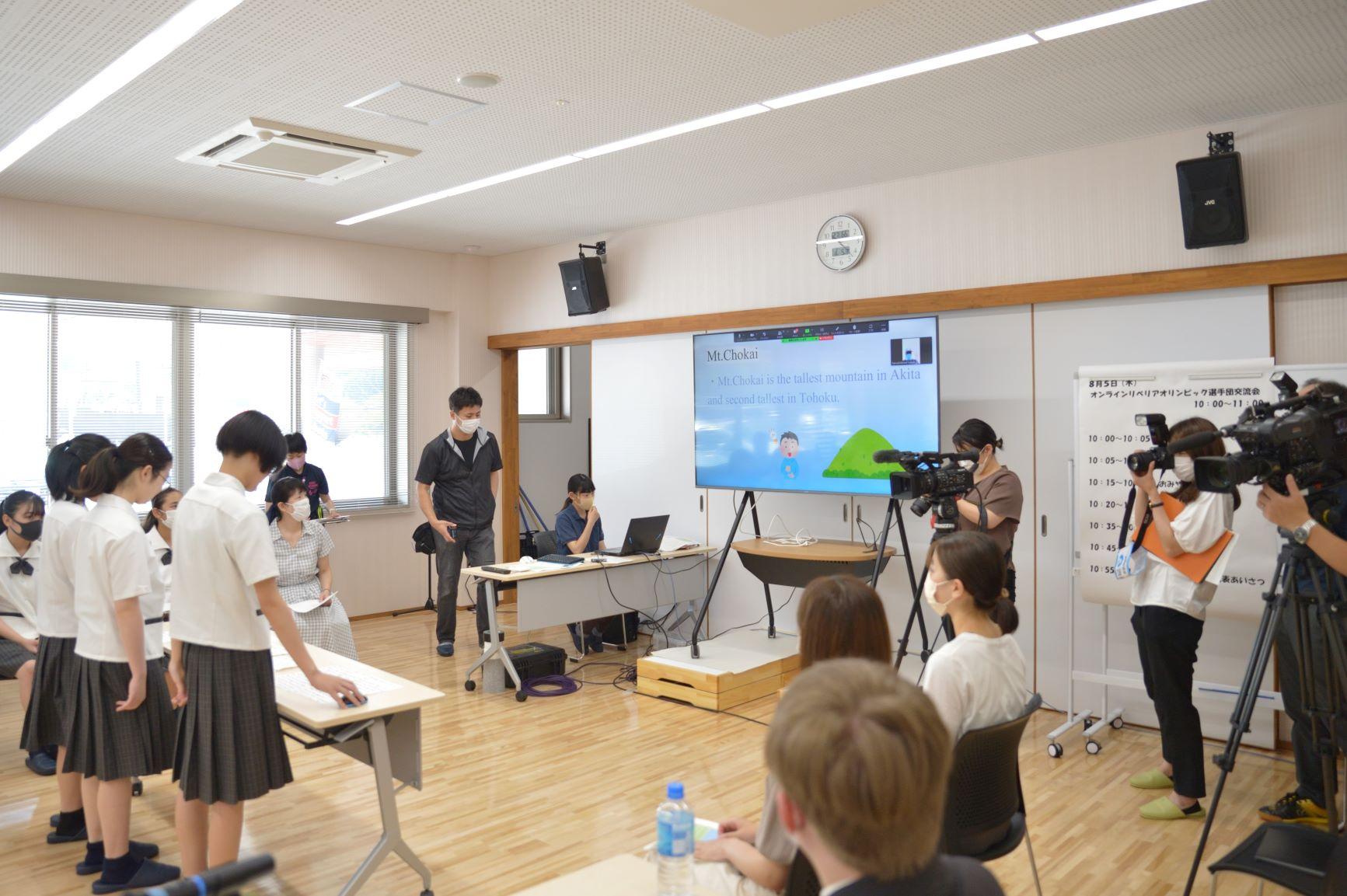 多目的屋内運動場内の会議室における、オンライン交流会の撮影風景の写真。室内にはカメラやディスプレイ等のオンライン用設備が配置されており、写真では丁度仁賀保高校の生徒たちが撮影機器の前で鳥海山について紹介する様子が収められている
