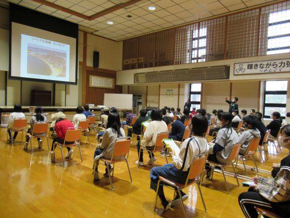 平沢小学校におけるリベリア講座の実施風景の写真。講座はスライドショー形式で進行されており、スクリーンにはリベリアの首都であるモンロビアの空撮写真が映し出されている