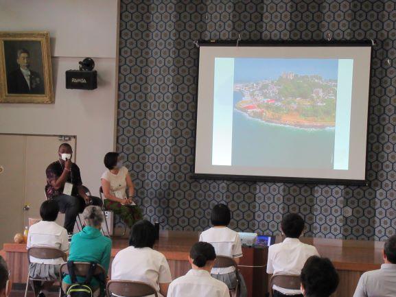 仁賀保公民館におけるリベリア講座の実施風景の写真。講座はスライドショー形式で進行され、壇上のスクリーンに資料映像を投影しつつ傍でスティーブンス・モール氏とジャイカの野口聡子女史が講演を行う様子が確認できる