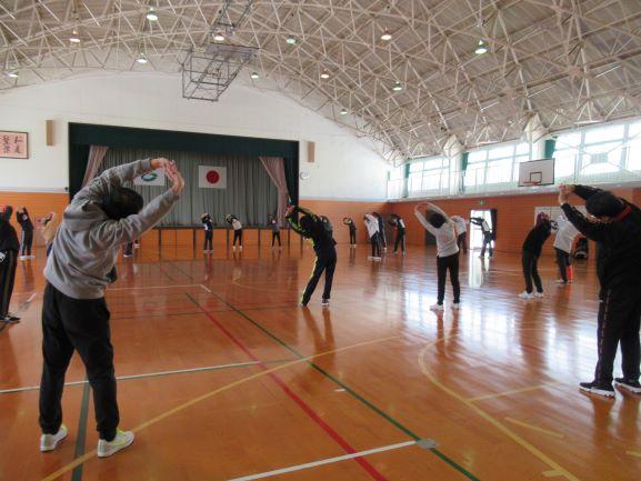 体育館にて、等間隔に並んでストレッチを行う参加者たちの写真。伸ばした両手を頭の上で組み、右や左に傾けている様子が確認できる