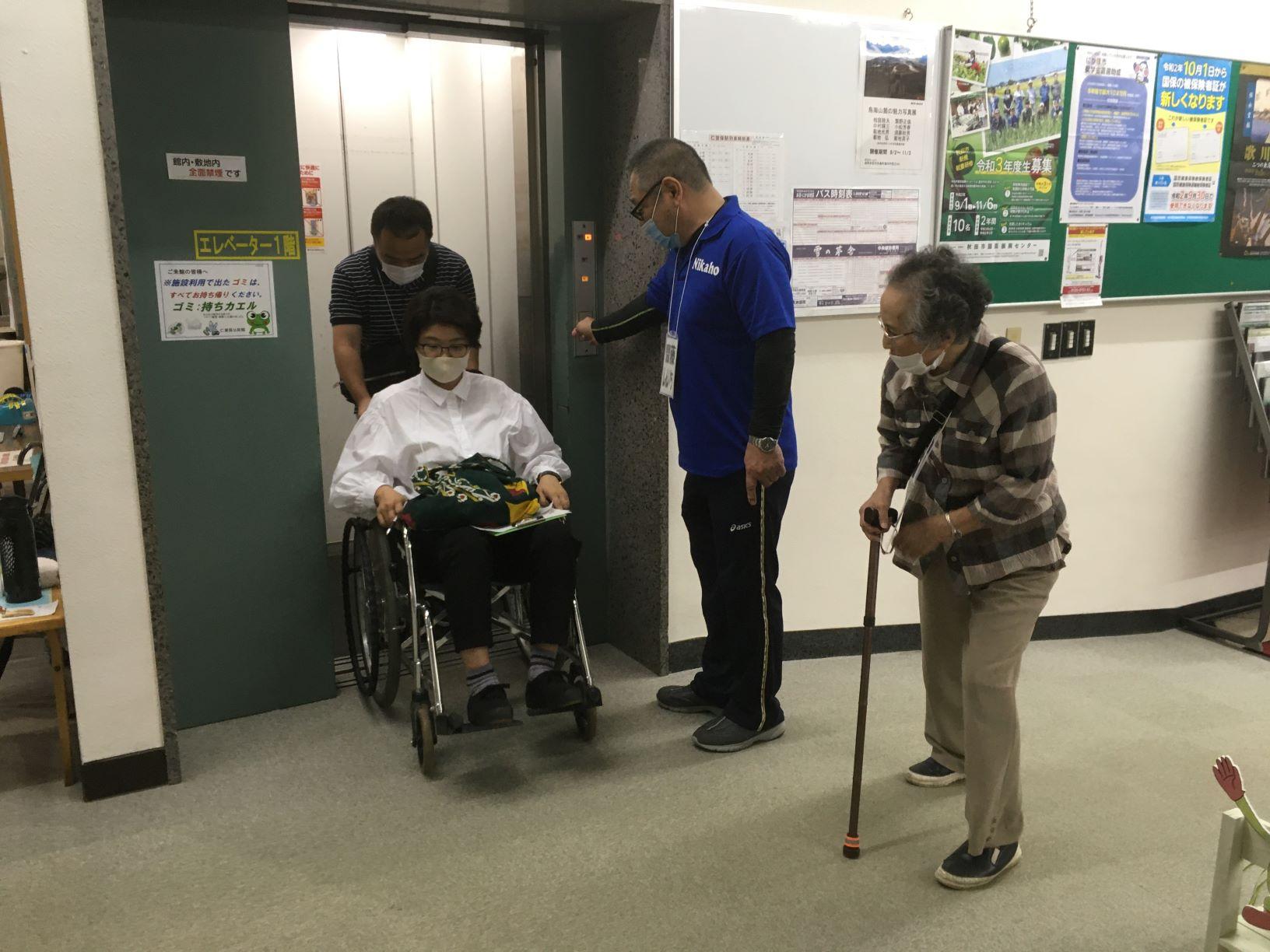 公民館1階のエレベーター前にて、実際に車椅子に乗ってエレベータを使用してみて問題点を確認する参加者たちの写真