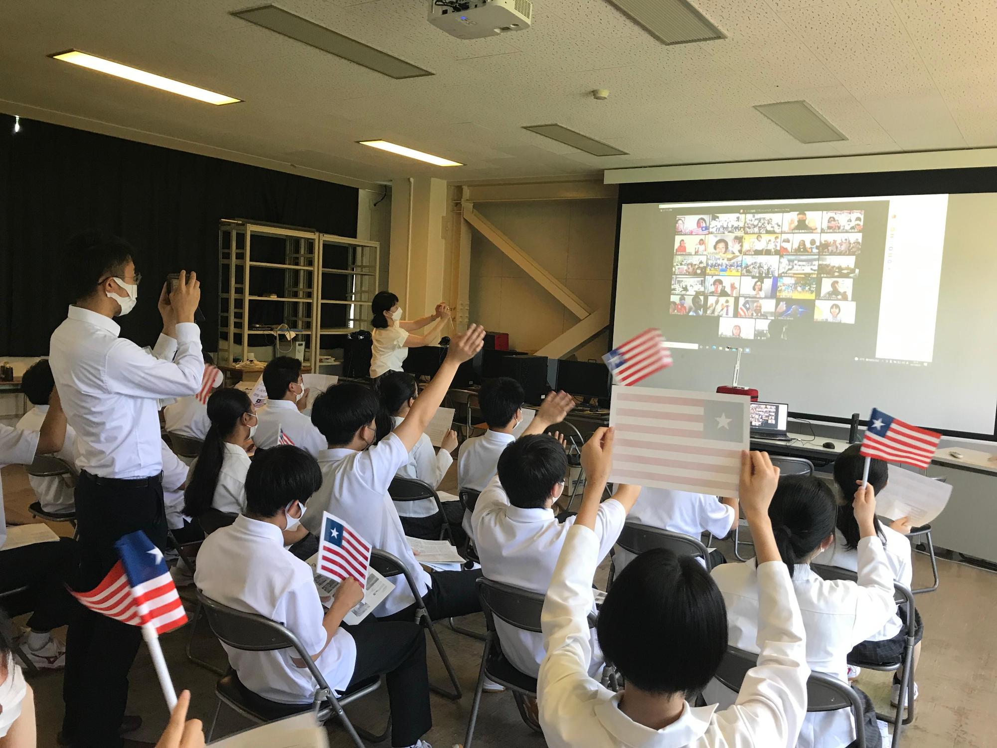 オンライン交流会に参加している最中の、仁賀保高校の視聴覚室を撮影した写真。スクリーン前の席に座った生徒たちがミニサイズのリベリア国旗を振る様子が確認できる