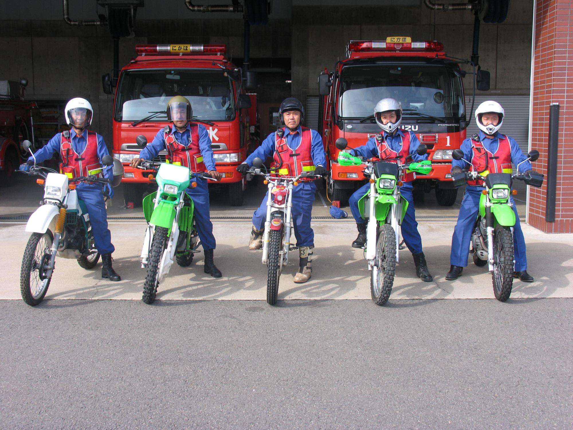 消防署の前でにかほ市消防団バイク隊の隊員5名が横一列に並んでバイクにまたがっている姿を正面から撮影した写真
