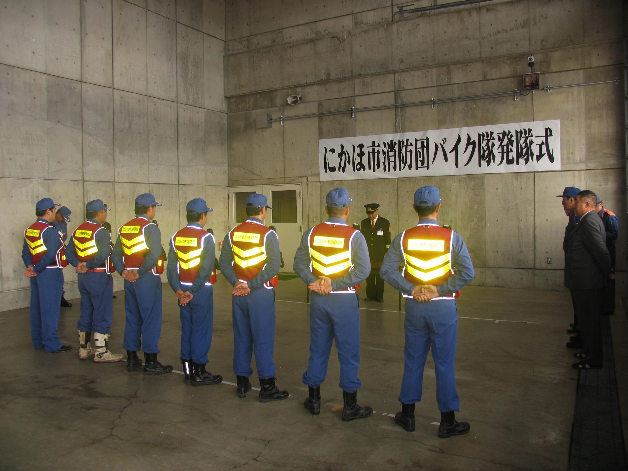にかほ市消防団バイク隊発隊式で横一列に並んだ7人の隊員たちが「休め」の姿勢で訓示を聞いている様子を後ろから撮影した写真