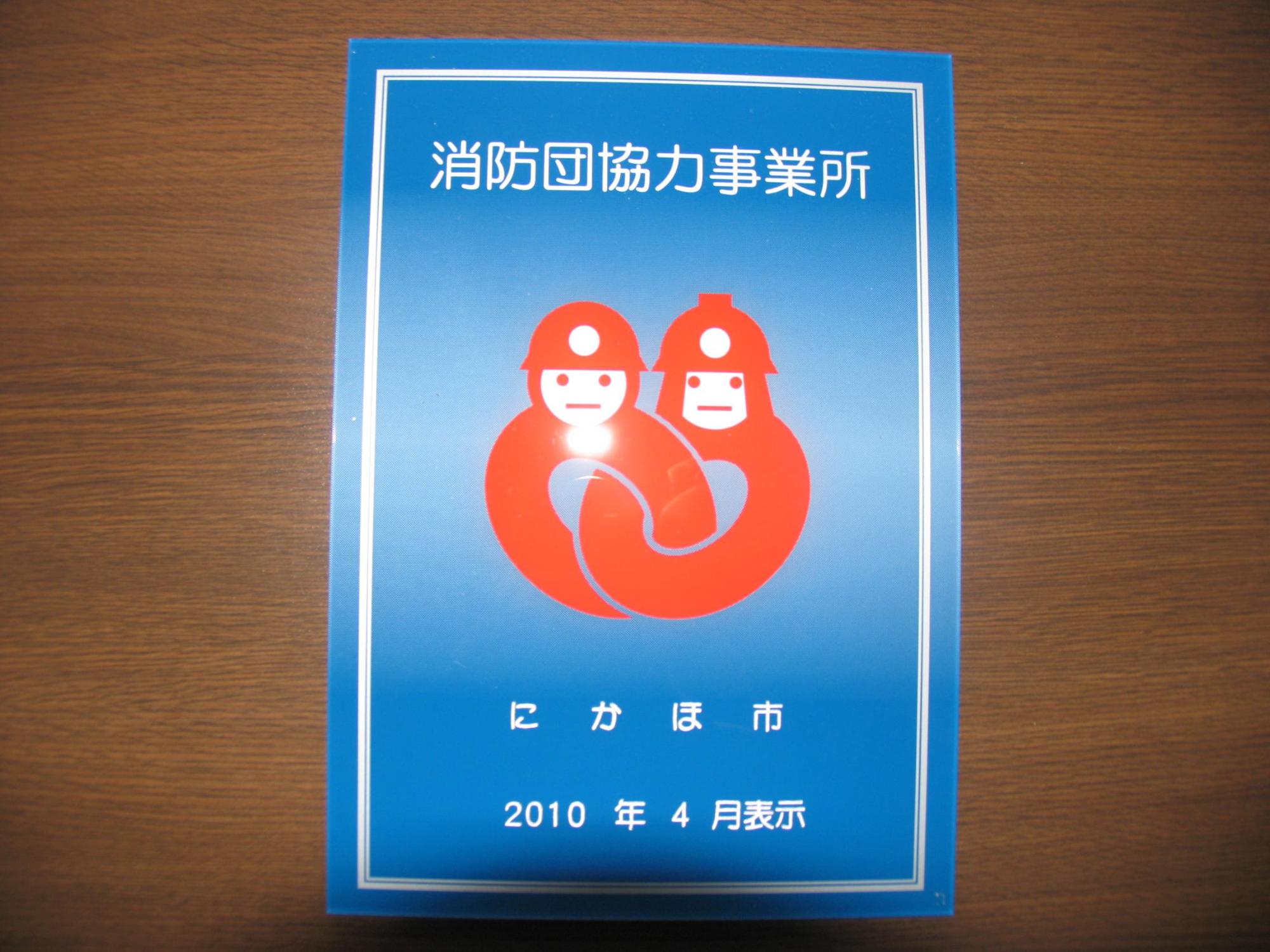 秋田県にかほ市が交付している青色の背景に赤色の消防団員のイラストが描かれた消防団協力事業所表示証を撮影した写真