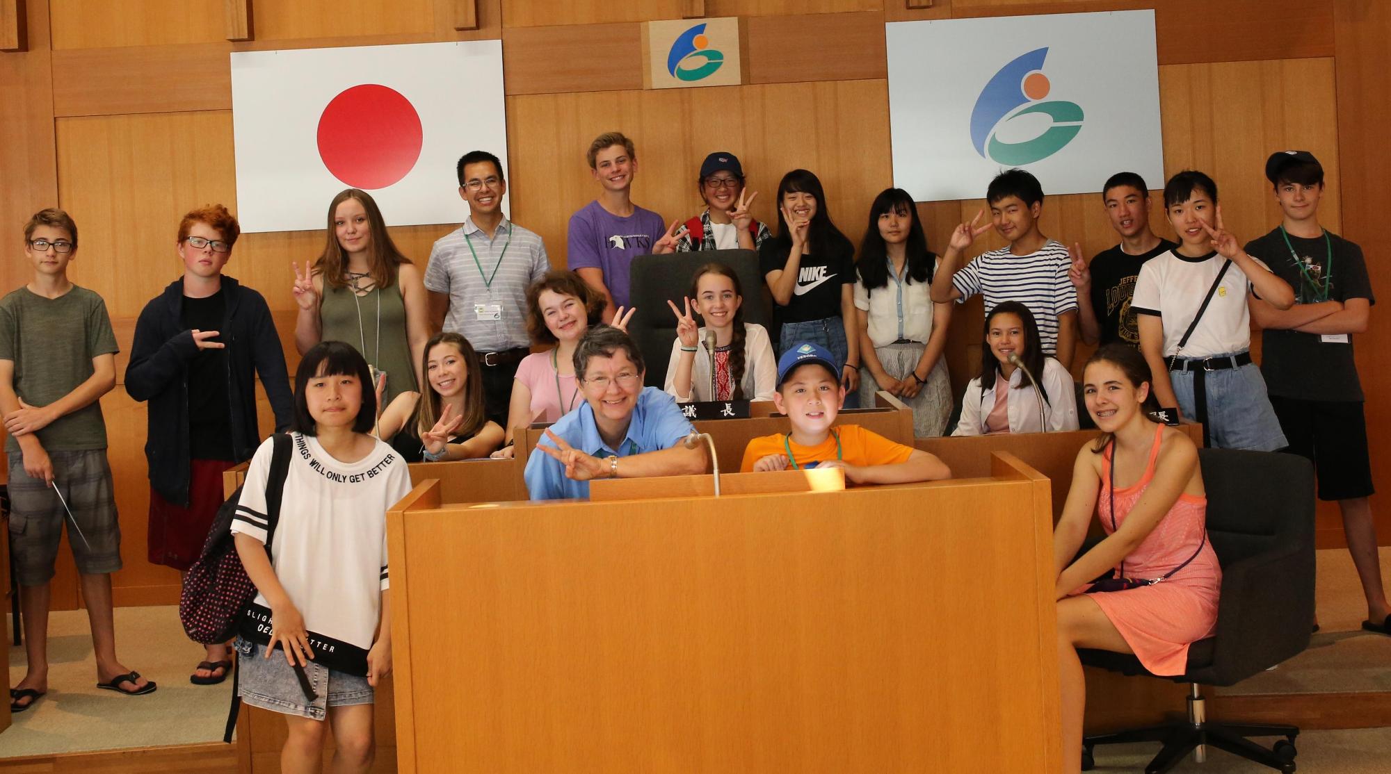 日本国旗とにかほ市のマークの旗が掲げられた室内で外国人を含む沢山の人達が3列に並んで笑っている集合写真