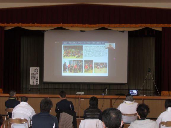 オンライン講演会の実施風景の写真。公民館内の舞台上にはスクリーンが設けられ、そこに投影された資料映像を参加者たちが視聴している様子が確認できる