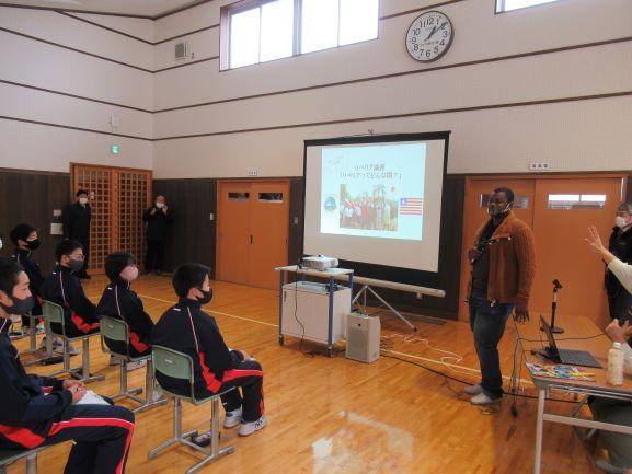 金浦中学校の屋内施設にて、壁際のスクリーンに映し出された映像を使って講演を行うスティーブンス・モール氏の写真