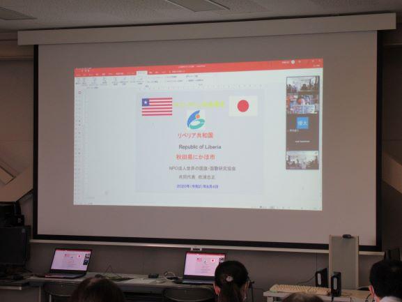 仁賀保高校の視聴覚室における「リベリアの国旗を学んでみよう」の実施風景の写真。講座はスライドショー形式で進行され、スクリーン上の映像では講座画面の右端で吹浦氏がオンラインで臨む様子が確認できる