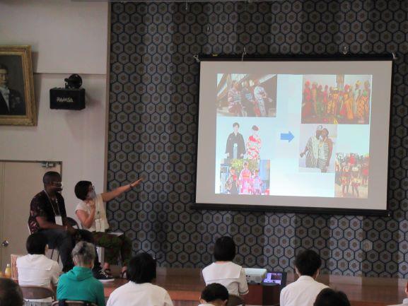 リベリア講座におけるスライドショーの一幕を収めた写真。スクリーンには、左側に日本の伝統衣装を着た人たちの写真3枚、右側にリベリアの民族衣装を着た人たちの写真3枚が映し出されている