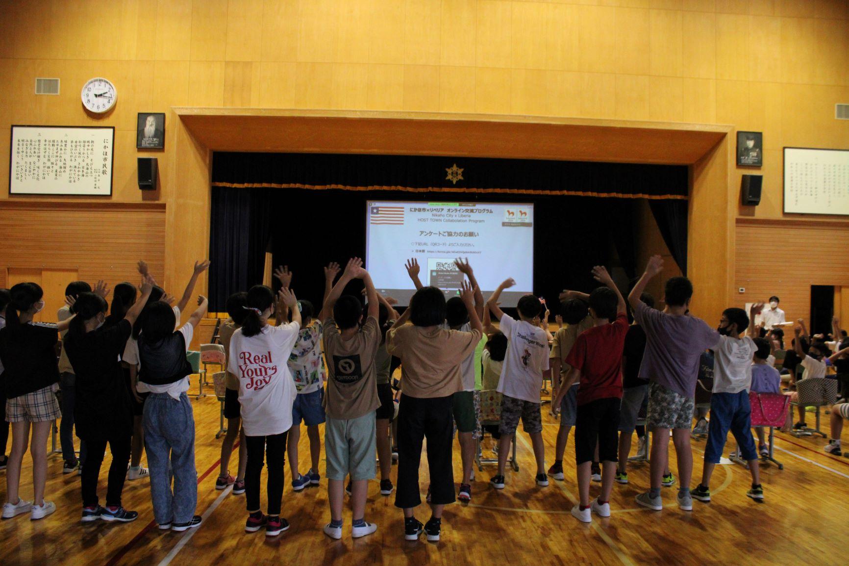 オンライン交流会に参加した金浦小学校の体育館にて、舞台のスクリーンに映し出された映像に手を振る児童たちの写真