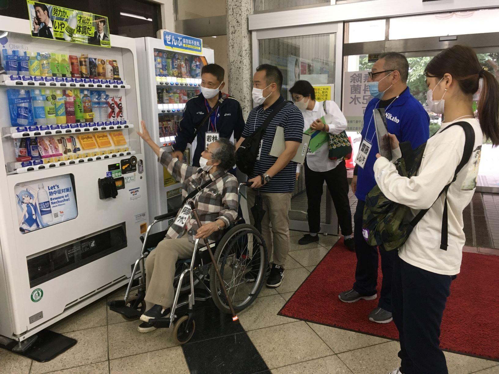 建物入口付近の自動販売機の前で、実際に車椅子に乗って自動販売機を利用する際の問題点を確認する参加者たちの写真