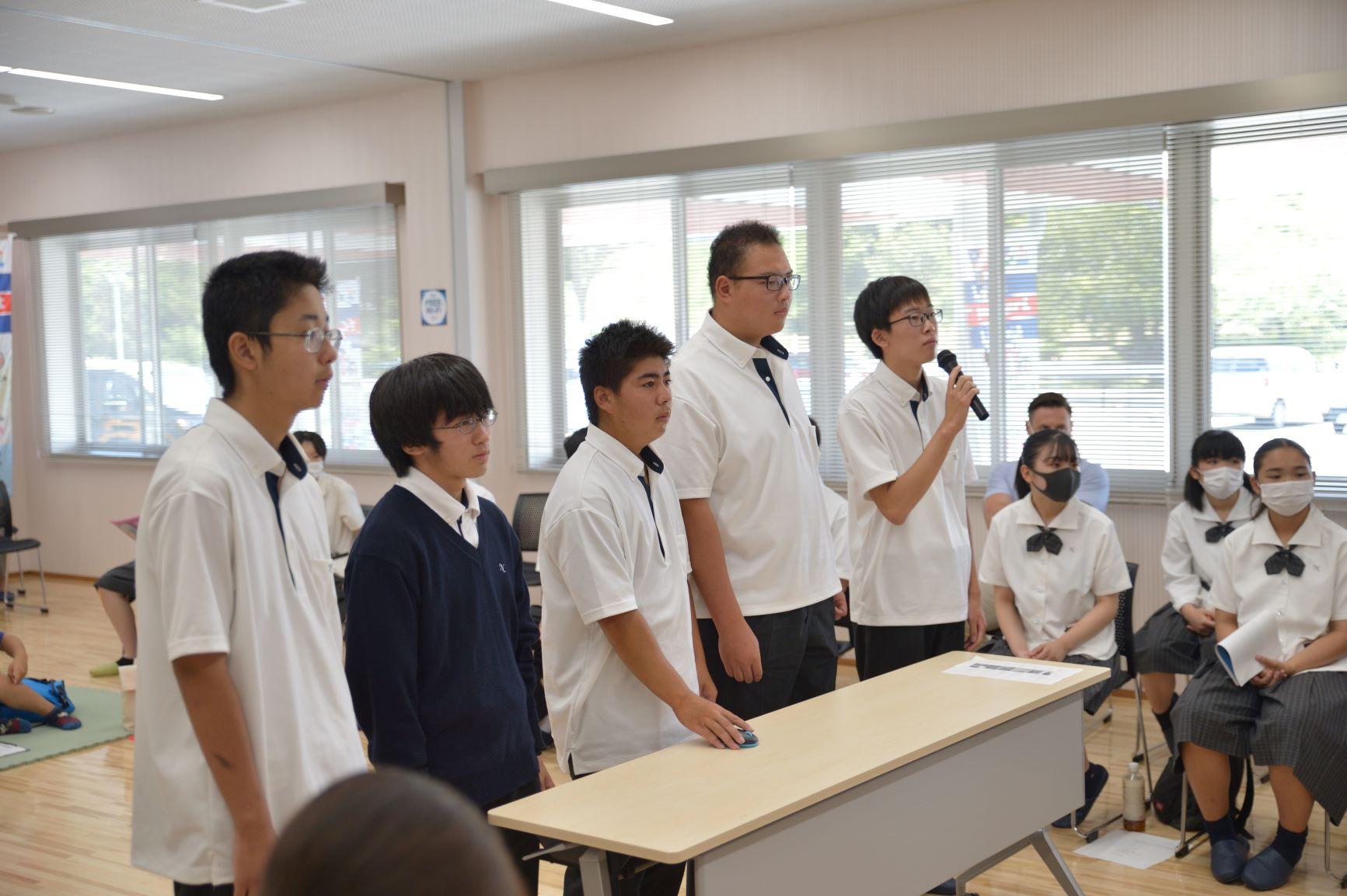 オンライン交流会にて、横一列に並んで交流に臨む仁賀保高校の男子生徒5名の写真。左端の生徒がマイクを持って話をし、中央の生徒が卓上のマウスを操作する様子が確認できる
