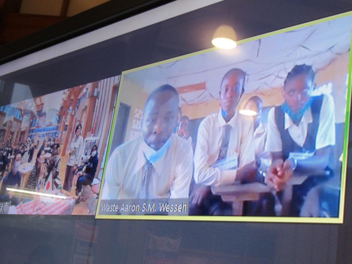 フードコート内の大型ディスプレイに映し出されたオンライントーク画面の写真。画面にはリベリアの高校生たちが映った映像と仁賀保市観光拠点センターでの様子の映った映像が左右に並んで分割表示されている様子が確認できる