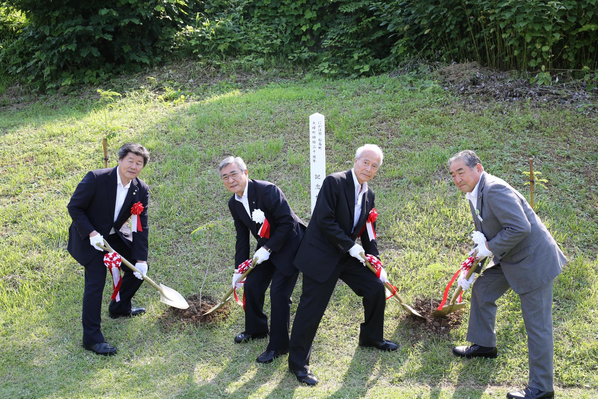 芝生が広がる場所でスーツ姿の4人の男性がシャベルを持って地面を掘っている写真