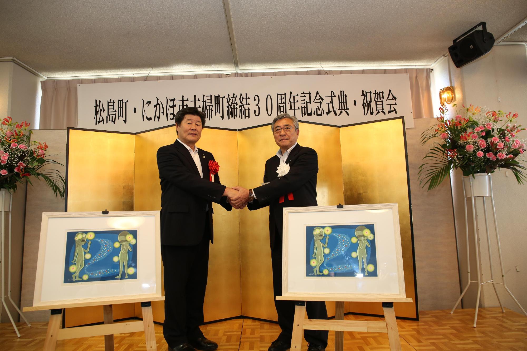 「松島町・にかほ市夫婦町締結30周年記念式典・祝賀会」と書かれた横断幕と金屏風をバックに、2人のスーツ姿の男性が両手で握手をしている写真