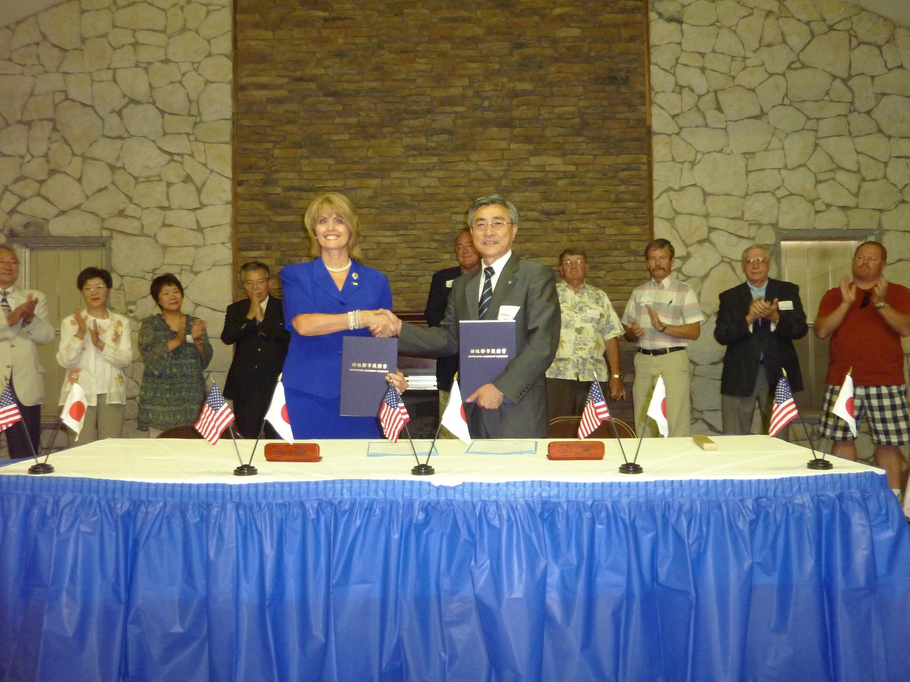 日本国旗とアメリカ国旗が交互に並べられた机の後ろで、男性と外国人女性が提携書を手に持ち握手し、それを見て沢山の人達が拍手をしている写真